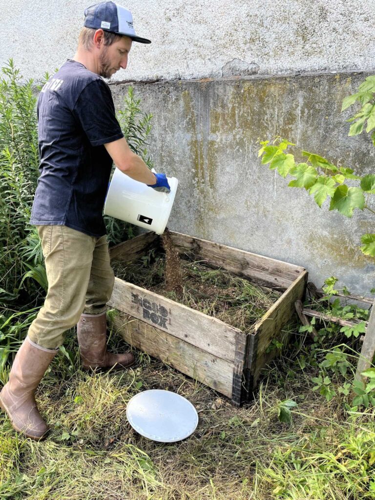 Mann leert den Inhalt einer Trockentoilette aus einem weißen Plastikkübel auf einen Komposthaufen