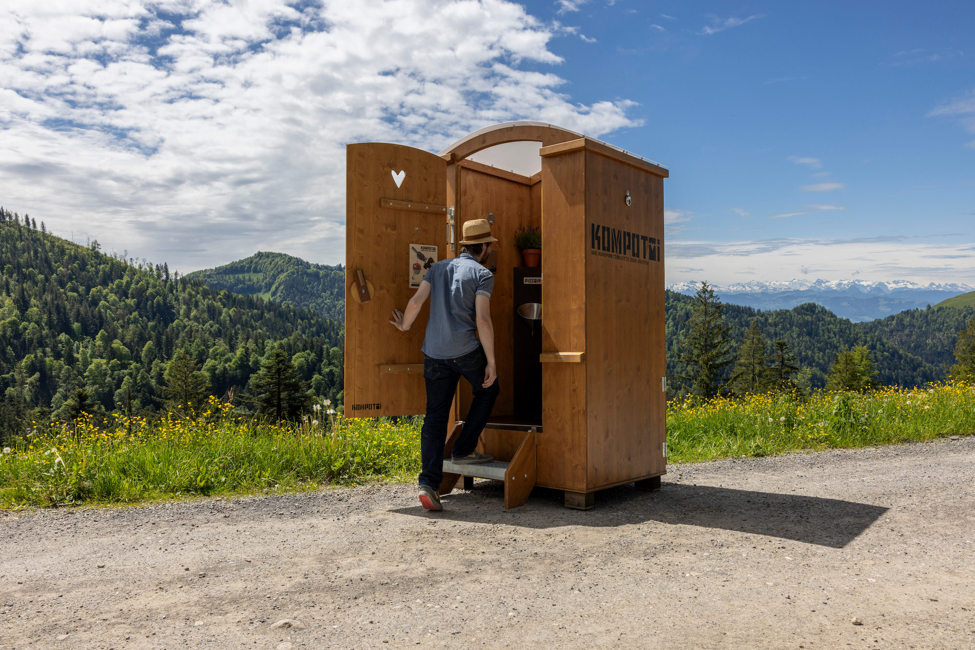 Vor einer Bergkulisse mit waldbedeckten Hügeln und Schneegebirge im Hintergrund geht ein Mann mit Strohhut grade in eine Kompotoi-Kabine aus Holz. Sie wirft einen Schatten auf den kiesigen Weg neben einer Wiese mit vielen gelben Blumen.