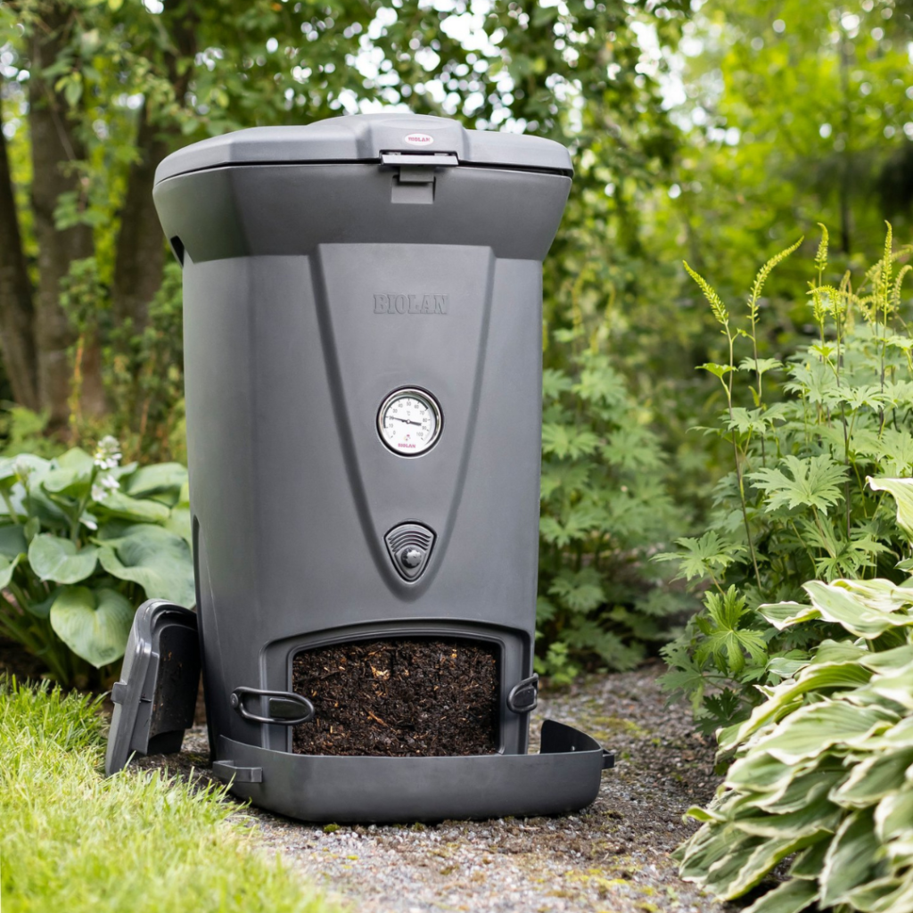 Der Biolan Thermo-Komposter 220eco steht mit geöffneter Entnahmeklappe unten am Gerät, aus der frische Komposterde schaut, in einem grünen Garten