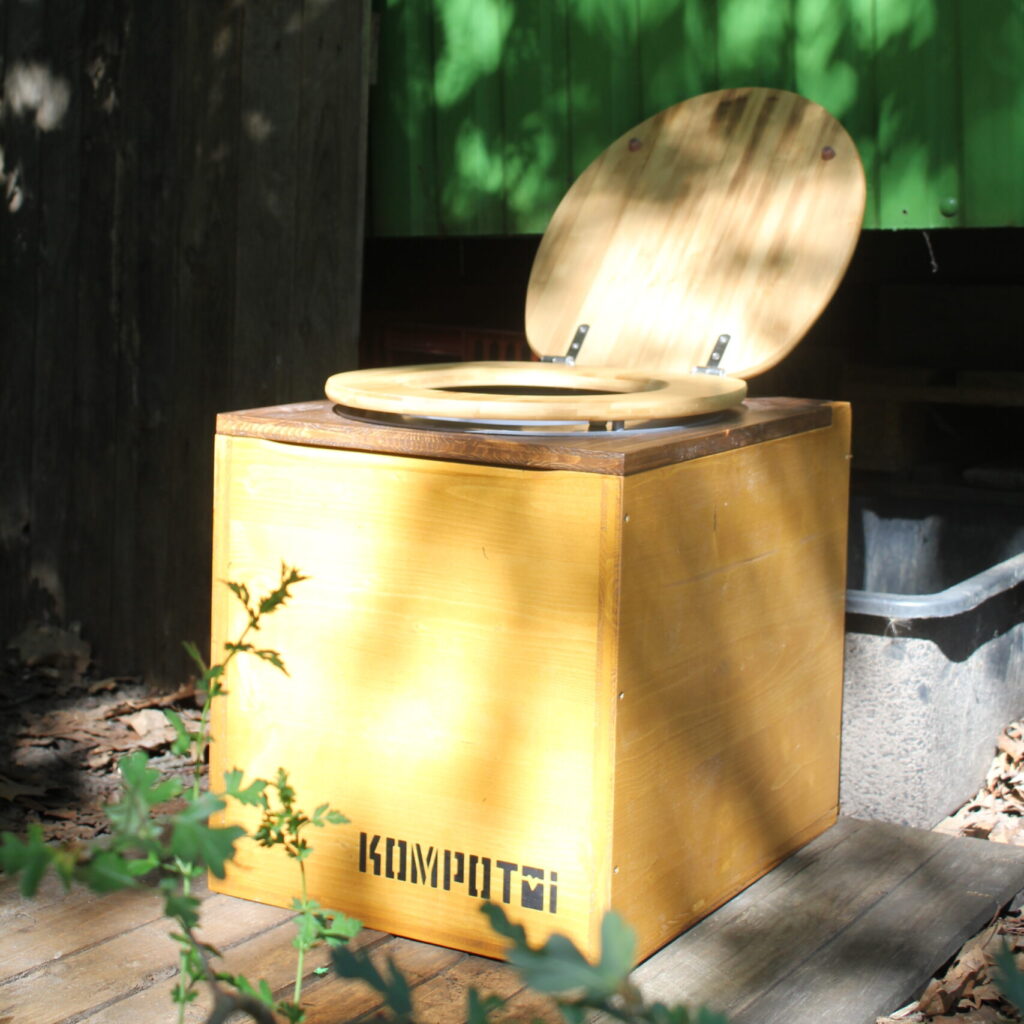 Kompotoi Petit steht mit Sonnenflecken beschienen im Garten hinter einem Bauwagen