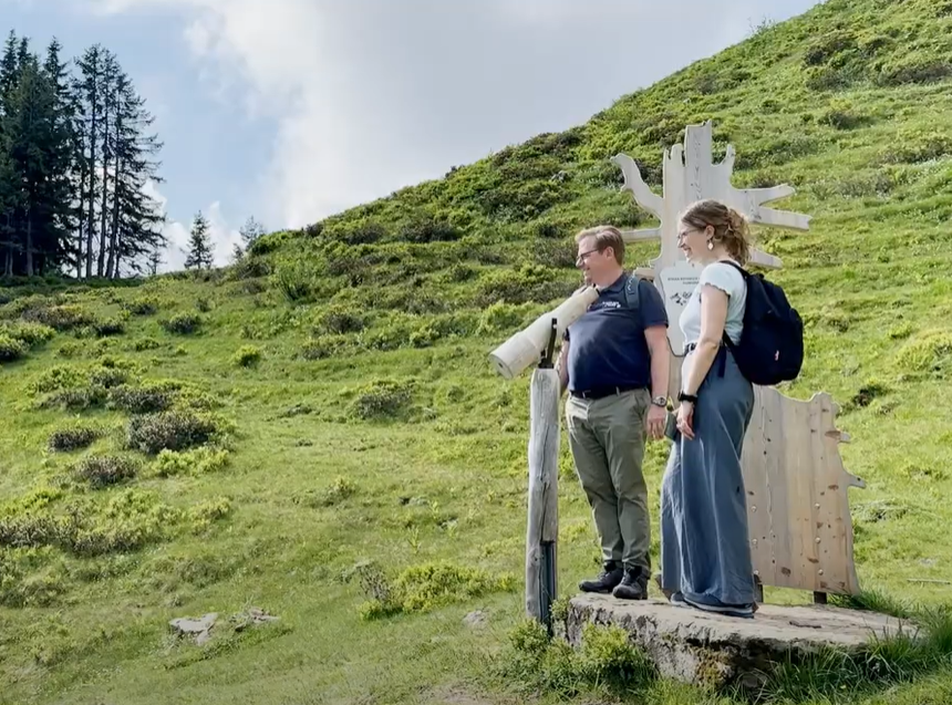 Grüne Bergkulisse, ein Mann und eine Frau stehen mit Rucksack rechts im Bild auf einer Holzplattform und schauen ins Tal, im Hintergrund dunkle Nadelbäume und strahlend blauer Himmel