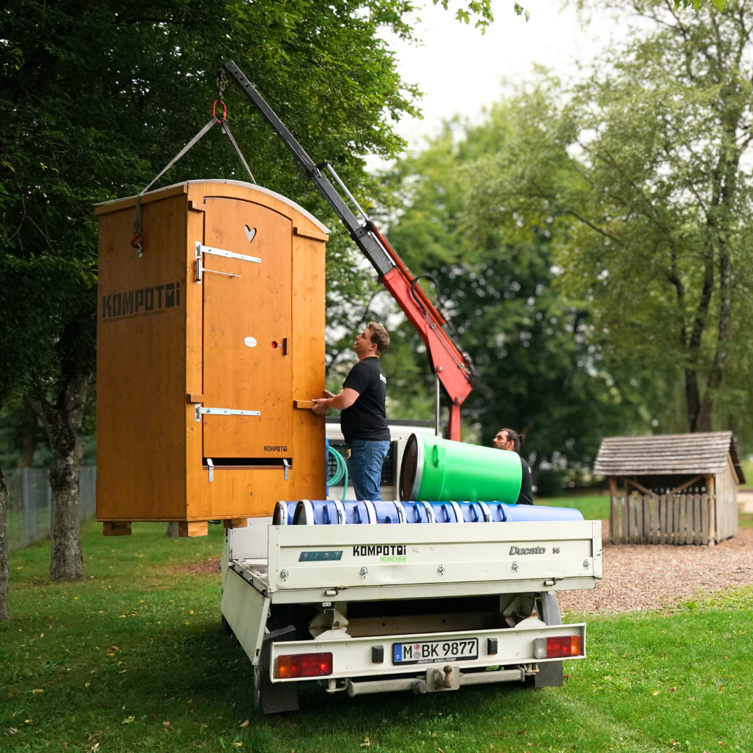 Mit einem kleinen Kran wird eine Holztoilette von Kompotoi vom Lieferwagen geladen. Ein Mitarbeiter der AWO schaut zu, um zu lernen. Im Hintergrund die Spielgeräte und Bäume.